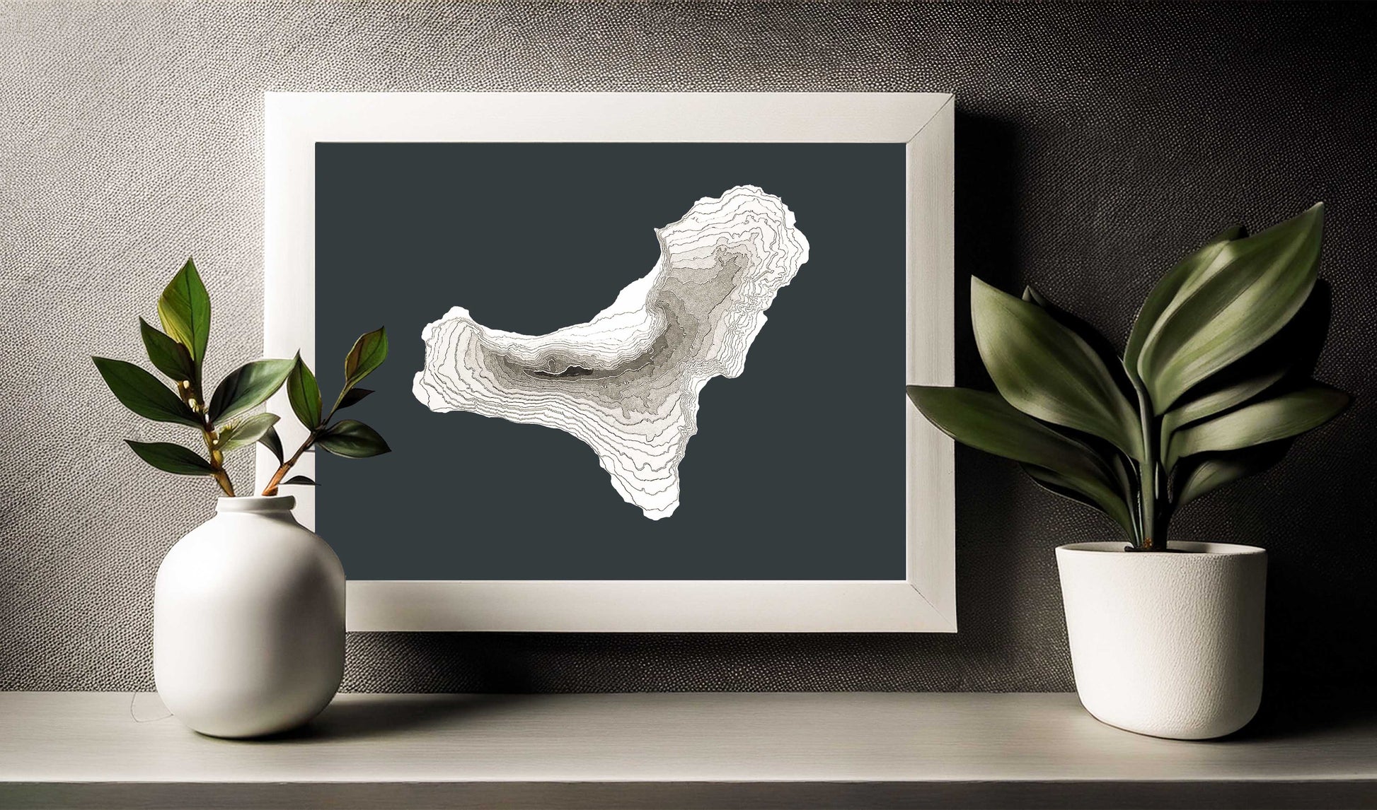 This is an image of the raised relief 3d map of El Hierro island in a  white wooden frame slightly above a shelf. Although the map is sold unframed it will look good in most kind of frames due to its greyscale colour palette.